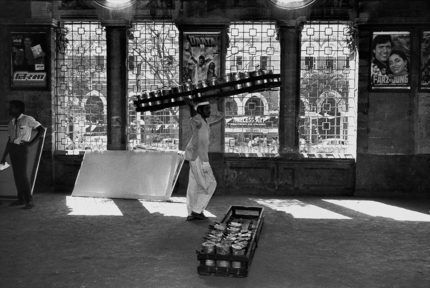 Black-and-white: a dabbawala walks through a shaft of hard window light carrying a long tiffin rack on his shoulder; a second rack rests on the floor; grilled arched windows and Bollywood film posters line the wall behind, a man leaning and watching at left